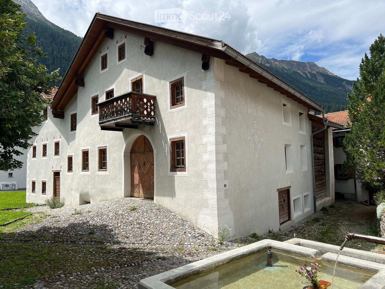 Large white building with brown roof, 2 floors, many windows, wooden door, wooden balcony, gravel path, fountain