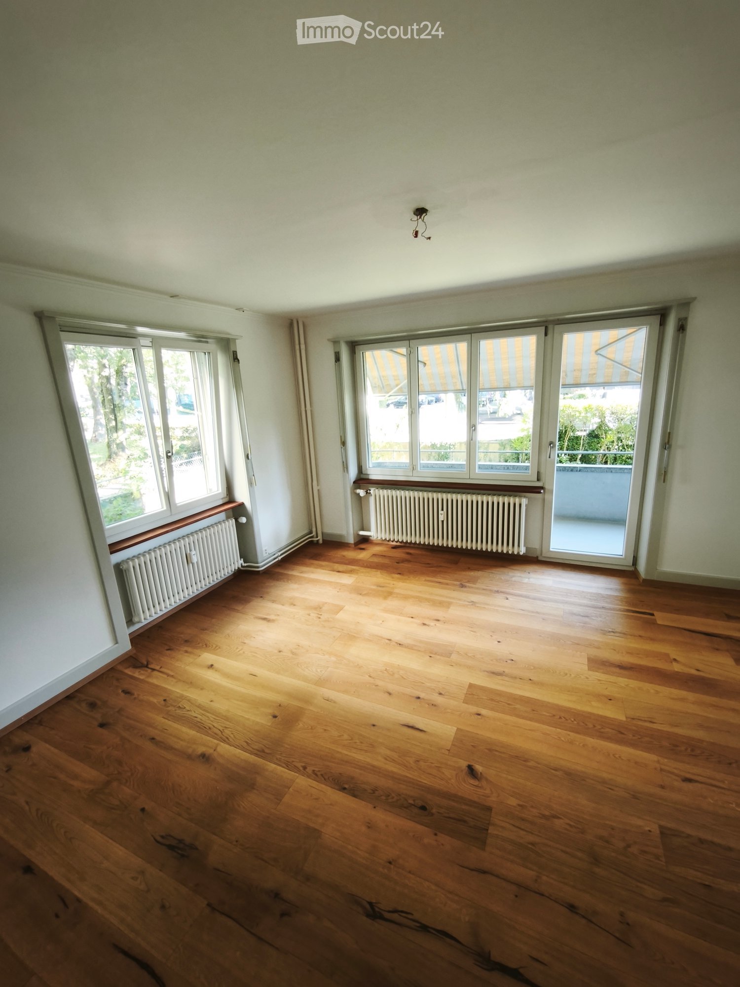 Empty room with wooden flooring, large windows, radiators, and a sliding glass door leading to a balcony.