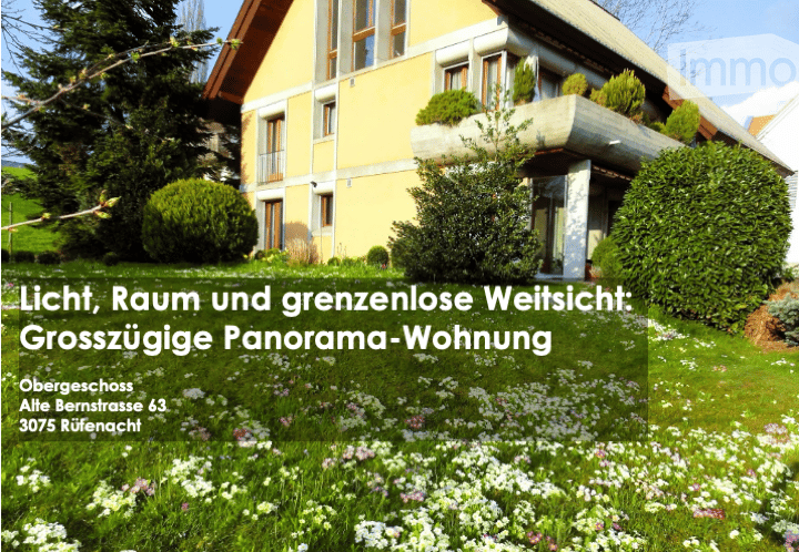 A two-story yellow house with white trim, multiple windows, and a balcony on the upper floor. The house is surrounded by a well-maintained garden with green bushes and flowering plants.