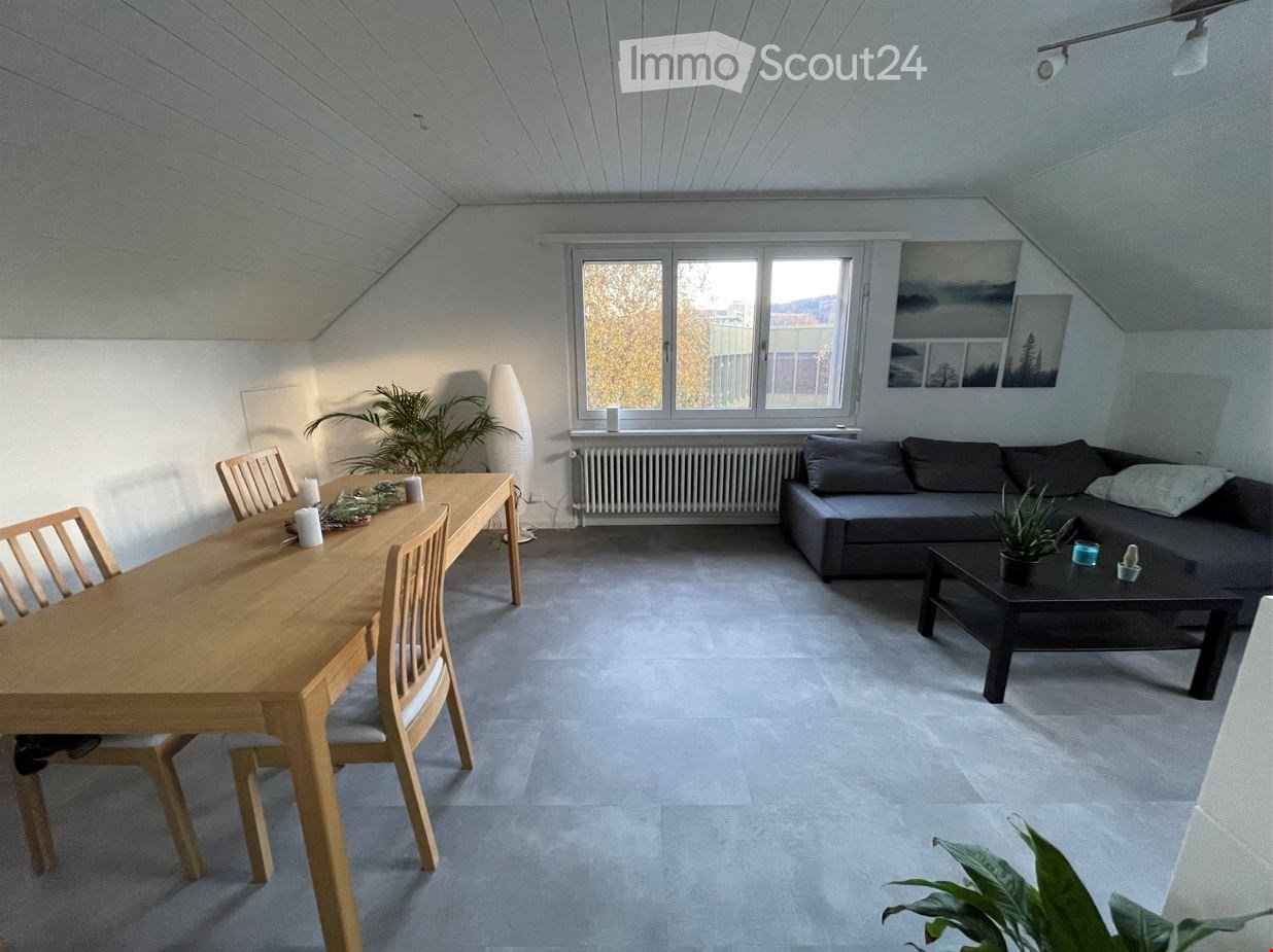 Bright living and dining area with grey sofa, coffee table, wooden dining table, chairs, and plants. Large windows provide natural light.