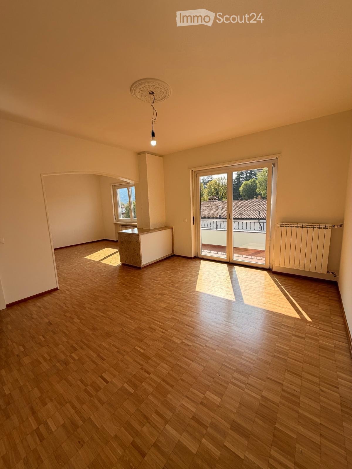 Empty room with wooden flooring, large sliding glass door leading to a balcony, and a small window on the left side.