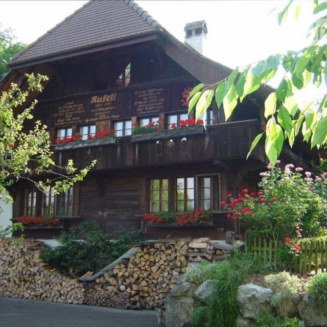 Traditional wooden house with a steep roof, multiple windows, and flower boxes on the balconies. The house is surrounded by greenery and a stack of firewood is visible in front.
