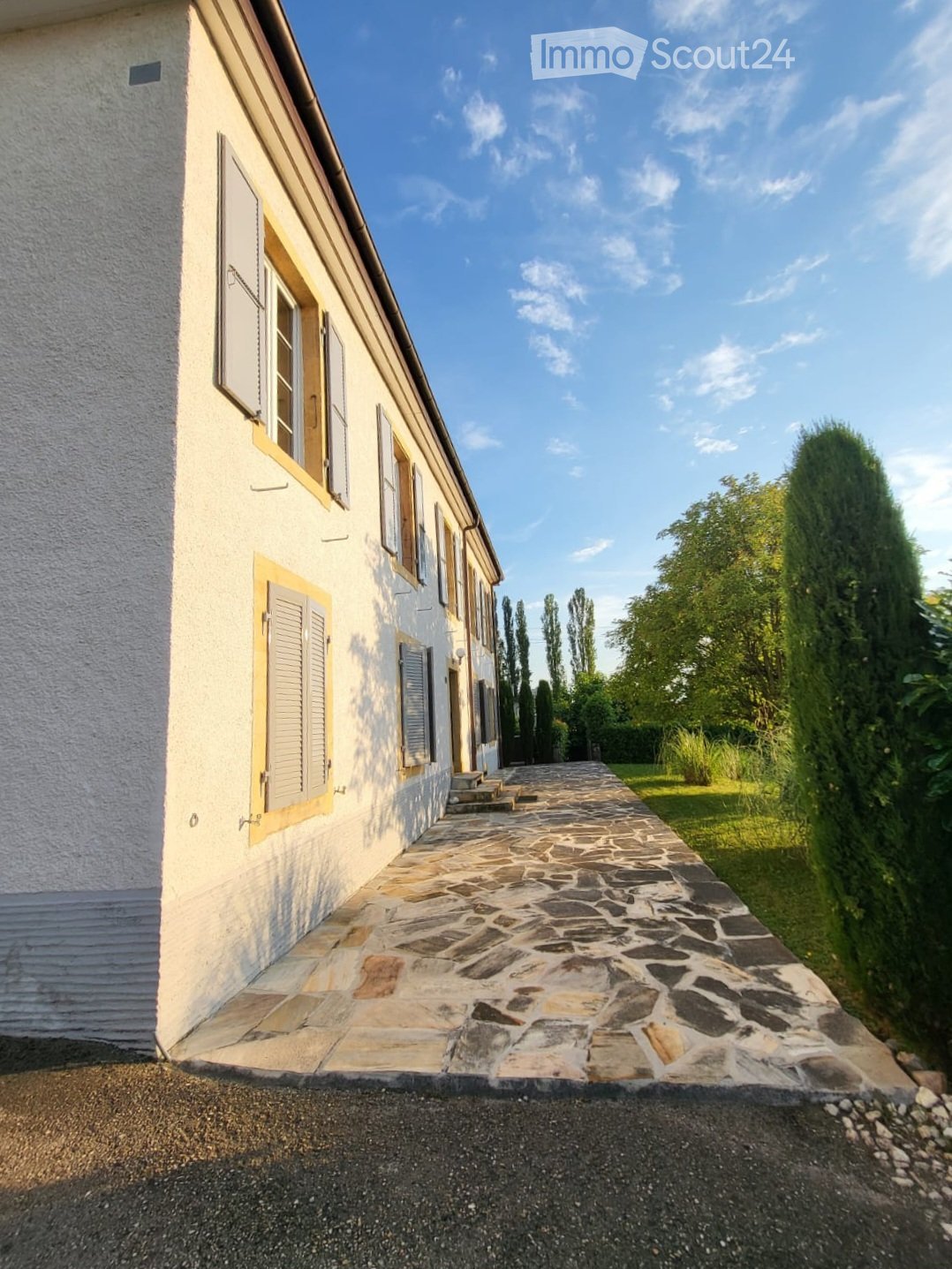 A long, single-story building with white walls and yellow-framed windows, featuring a stone-paved pathway leading to the entrance.