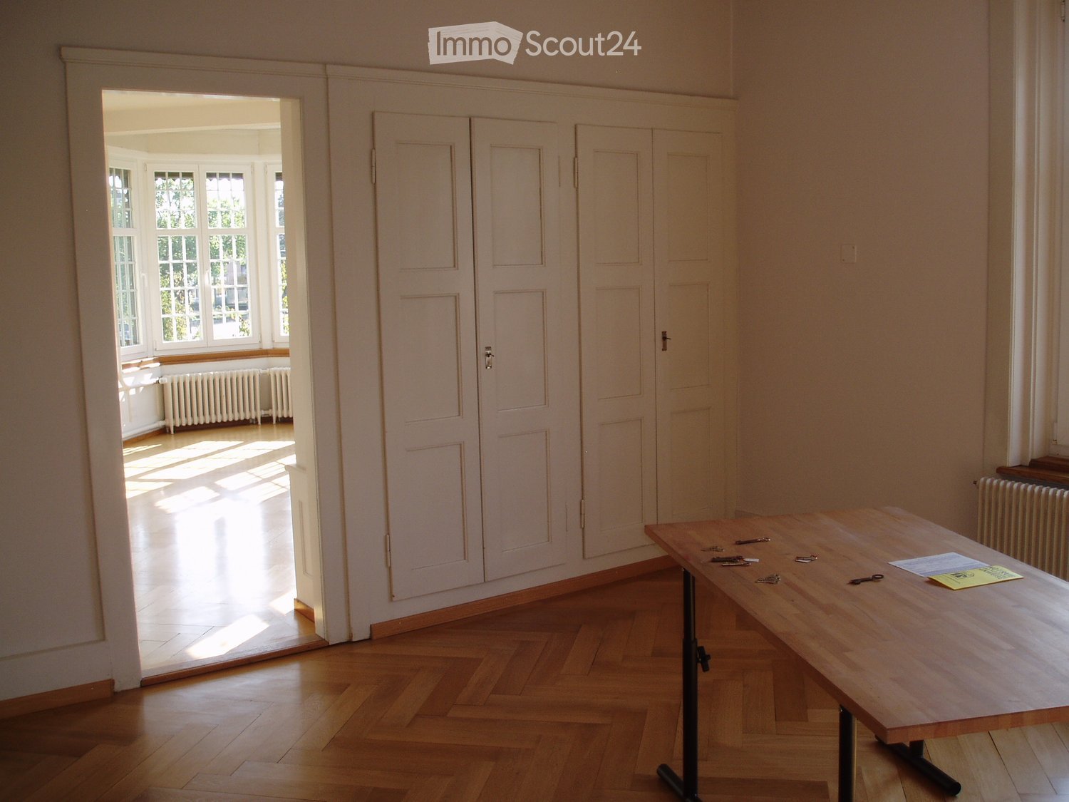 Bright room with wooden floor, large double doors, radiator, and a wooden table with keys and papers on it.