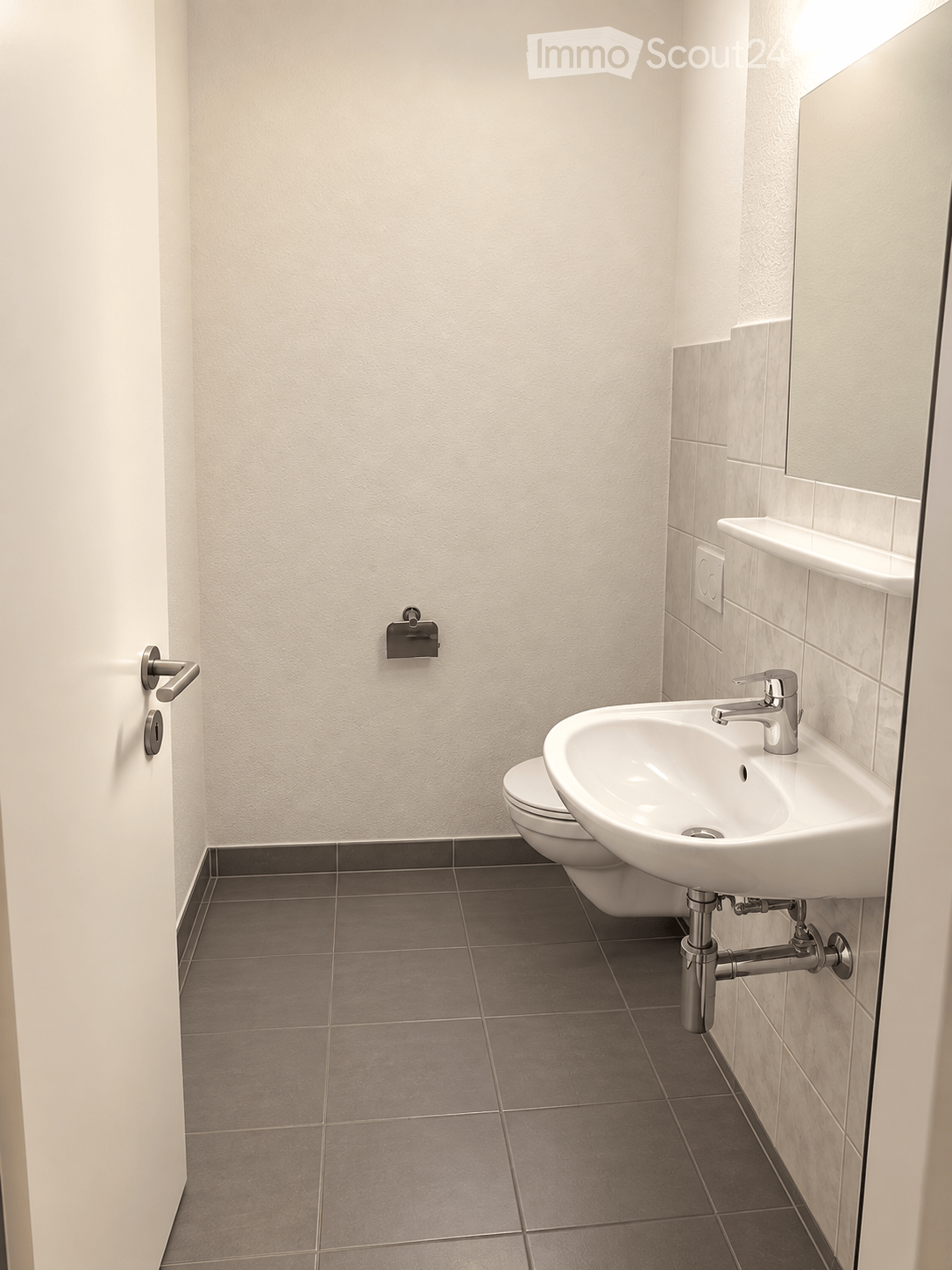 Modern bathroom with a white sink, chrome faucet, toilet, and a mirror above the sink. The walls are tiled in light beige, and the floor is covered with dark gray tiles.
