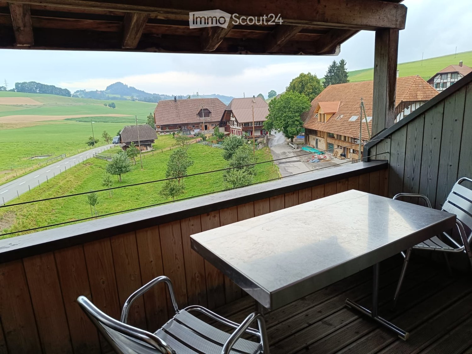 Balcony with wooden railing, metal table, and chairs, overlooking a scenic countryside with houses and green fields.