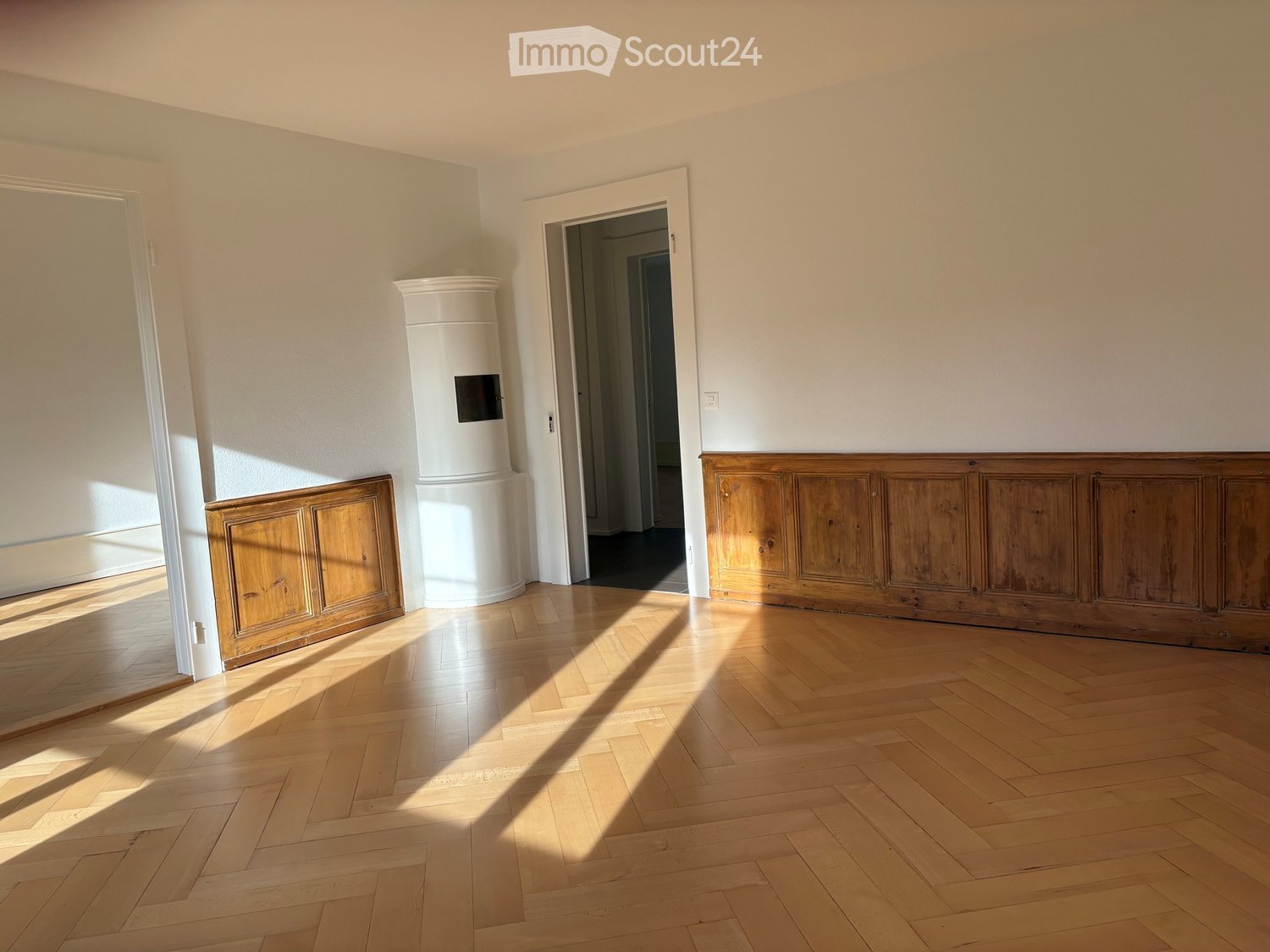 Empty room with wooden herringbone flooring, white walls, and a white cylindrical object in the center. Two doorways lead to adjacent spaces.