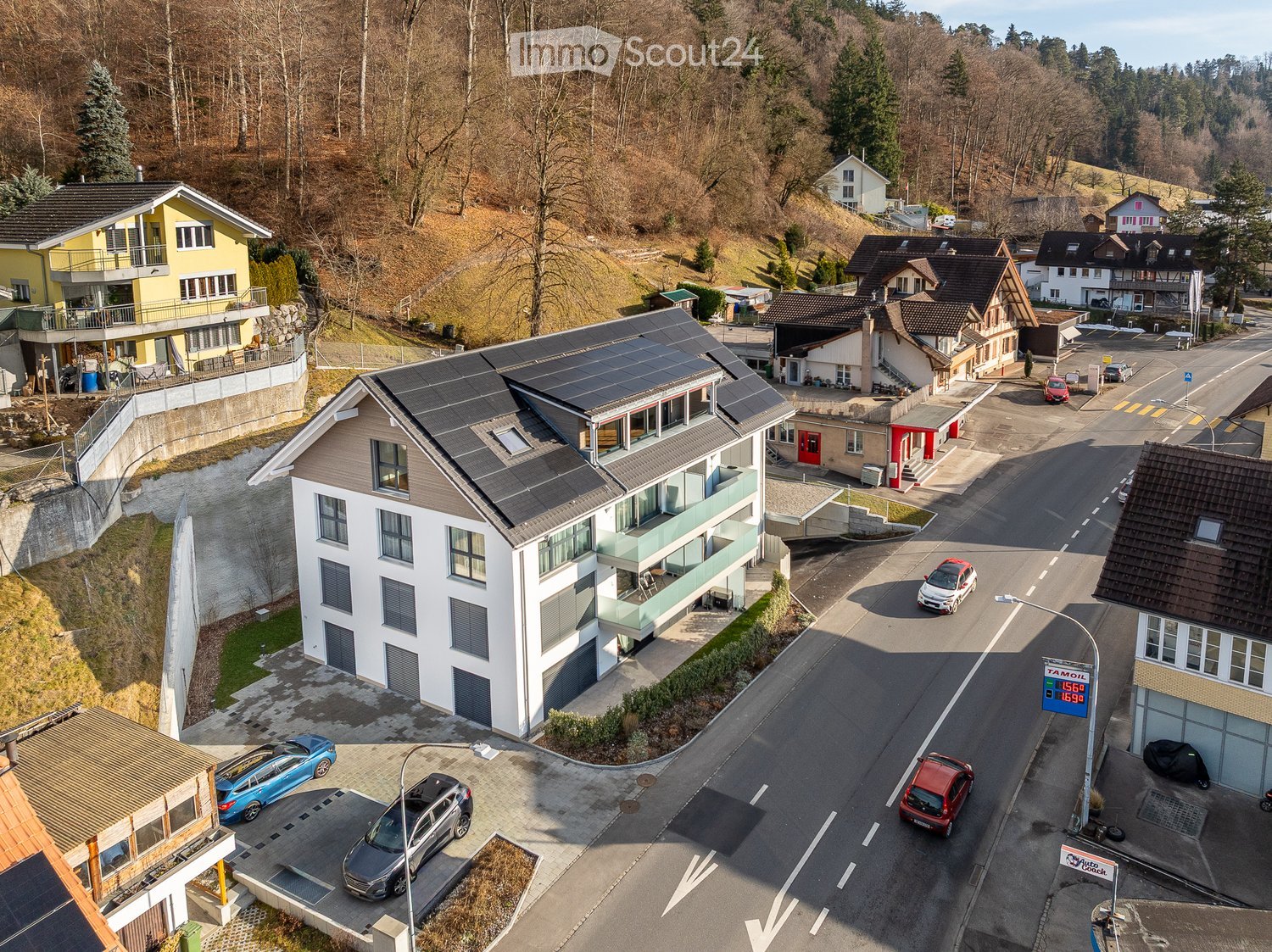 Aerial view of a residential area with multiple houses, solar panels on roofs, parked cars, and a road running through the neighborhood.