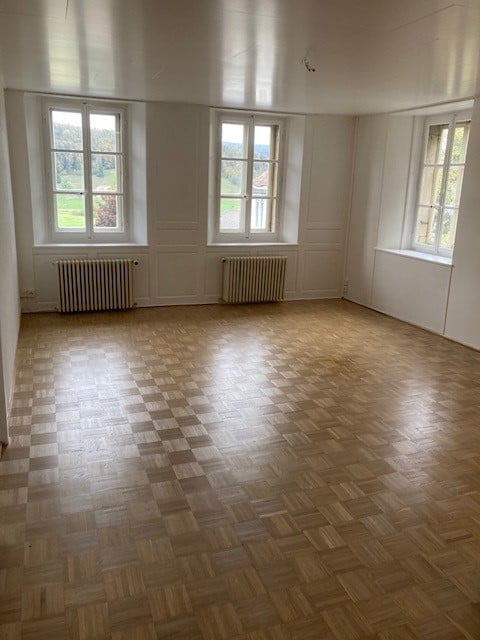 Empty room with wooden parquet flooring, white walls, and three large windows offering a view of a green landscape. Radiators are visible under the windows.