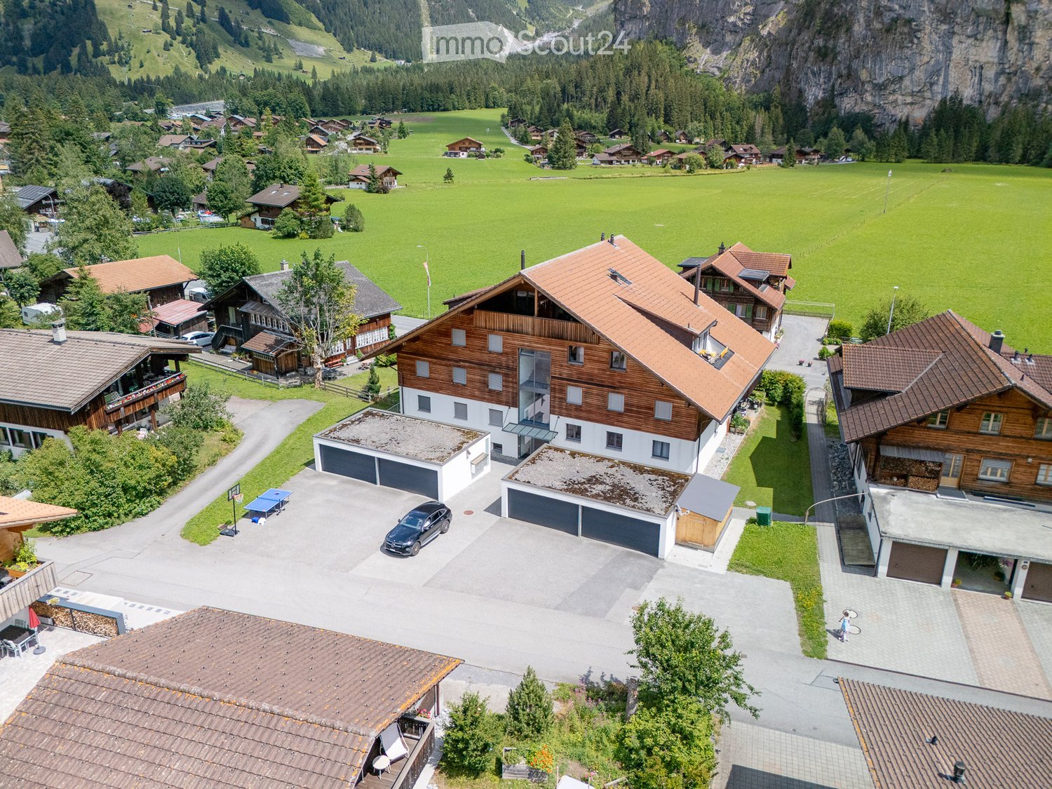 Aerial view of a rural village with traditional wooden houses, a large central house with a garage, and surrounding green fields and mountains.