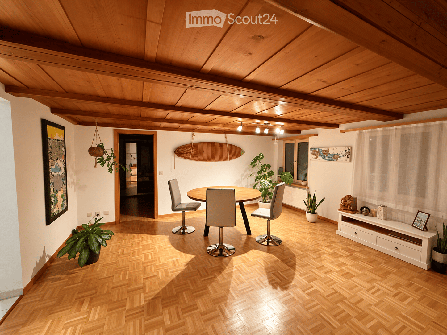 A modern dining area with a round wooden table, four chairs, potted plants, a shelf with decorative items, and a surfboard on the wall.