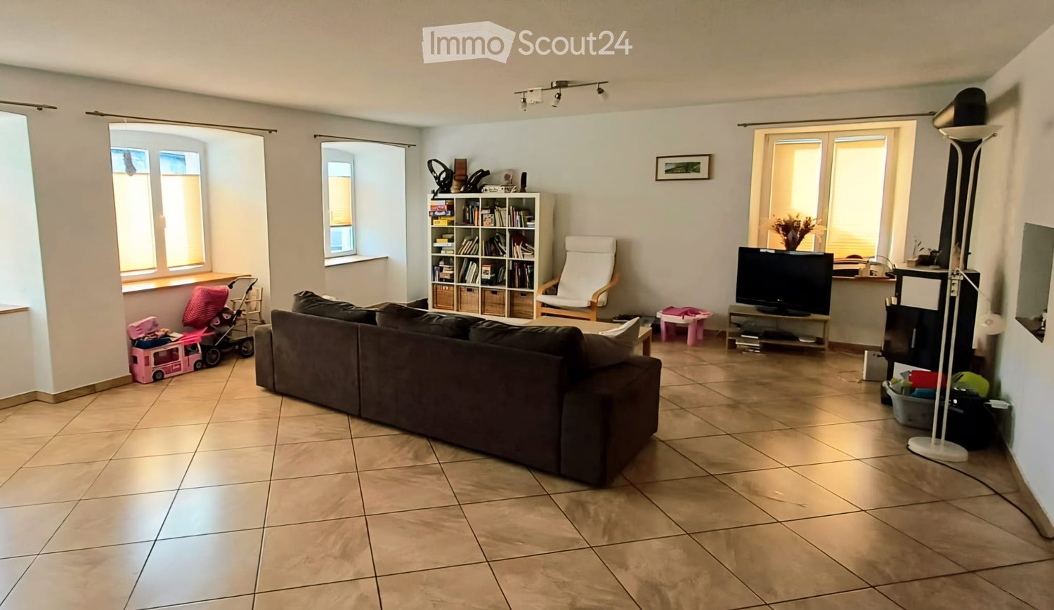 Bright living room with tiled floor, large windows, sofa, armchair, bookshelf, TV, and toys.