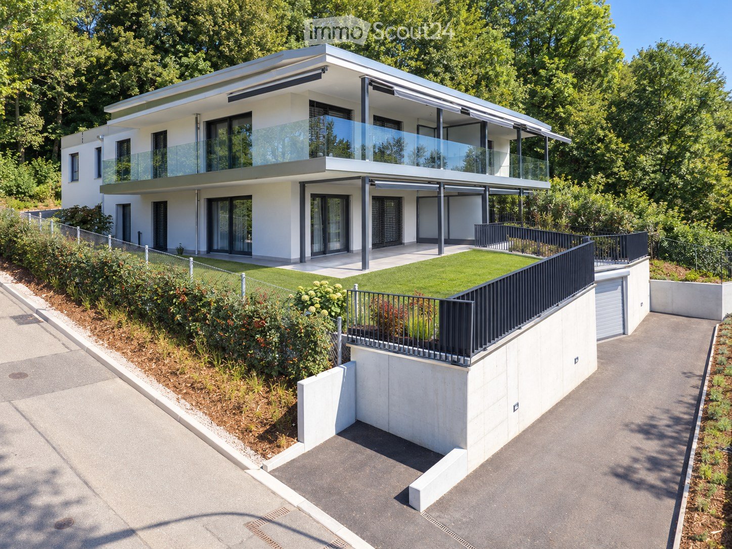 Modern two-story house with white exterior, large glass windows, and a raised ground floor. Features include a balcony, garage, and landscaped garden area.
