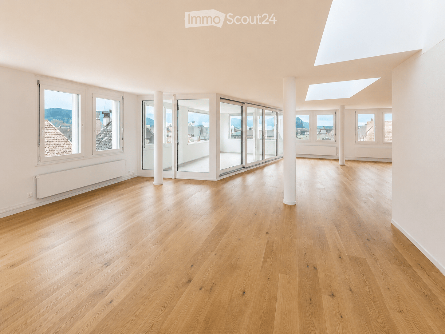 Bright, empty room with light wooden flooring, large windows, and sliding glass doors leading to a balcony. The space features white walls, a skylight, and a radiator on one wall.