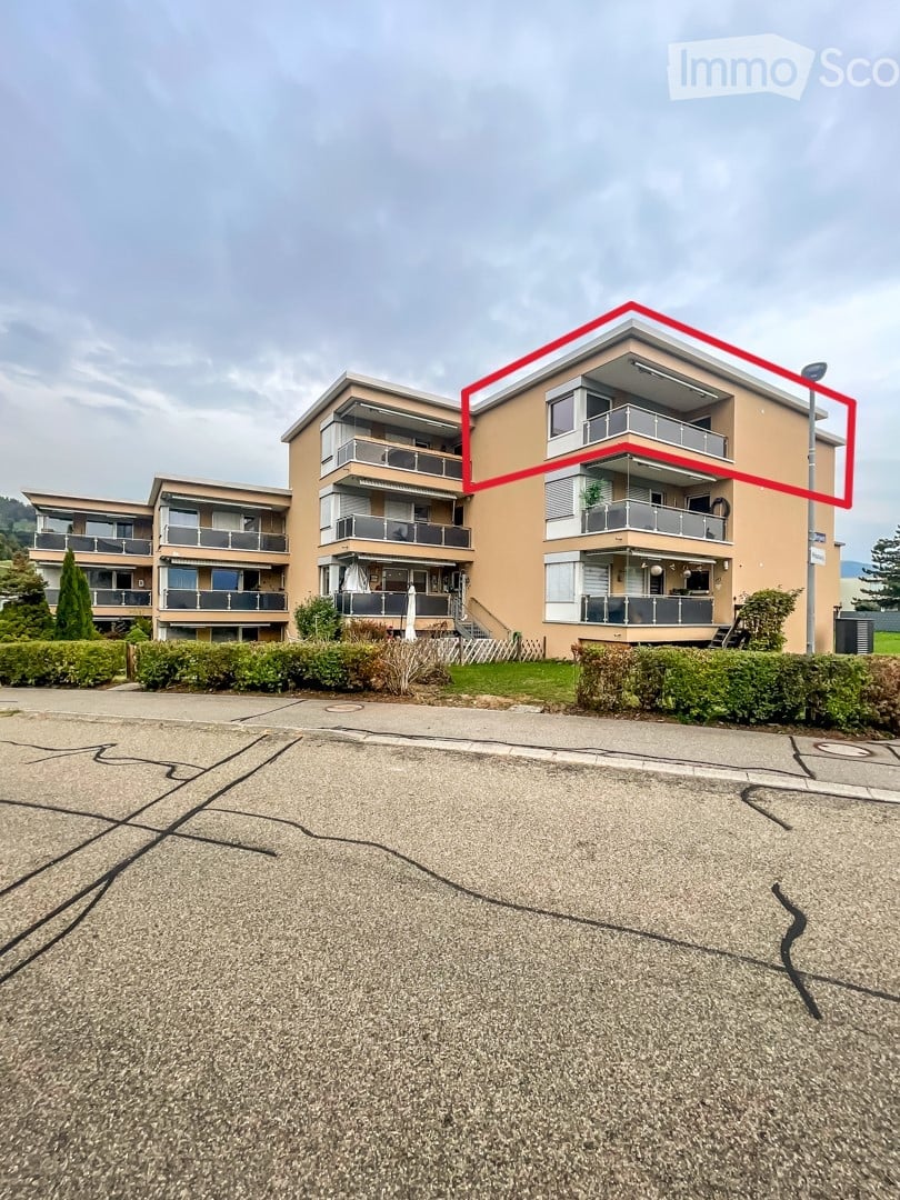 Multi-story residential building with balconies and modern design, surrounded by greenery and a paved parking area.