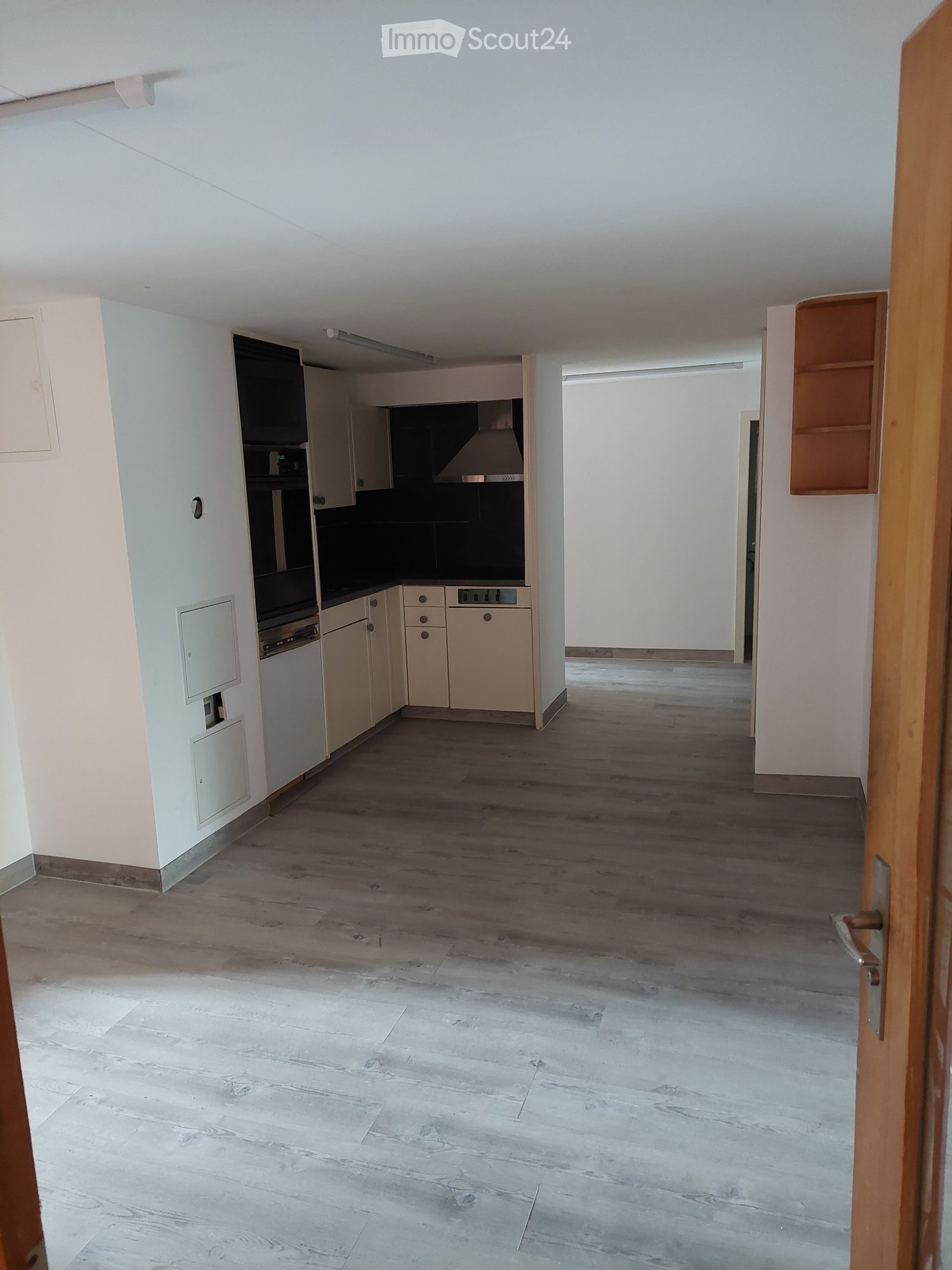 Empty room with light wooden flooring, white walls, and a small kitchen area with cabinets and appliances visible in the background.