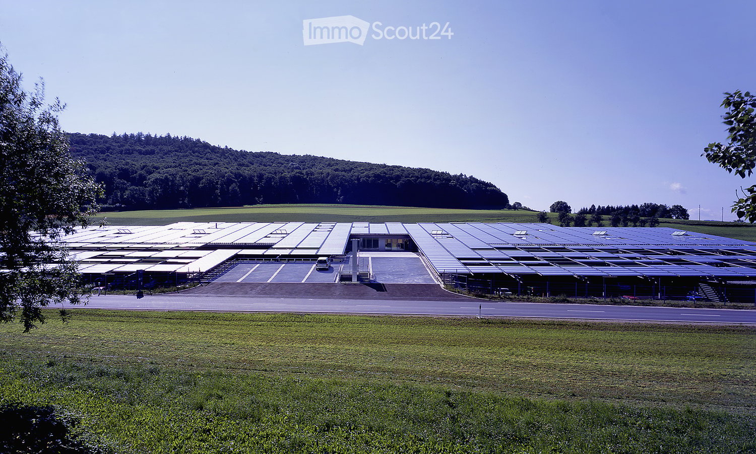 Large industrial building with solar panels on the roof, surrounded by open fields and a parking area.