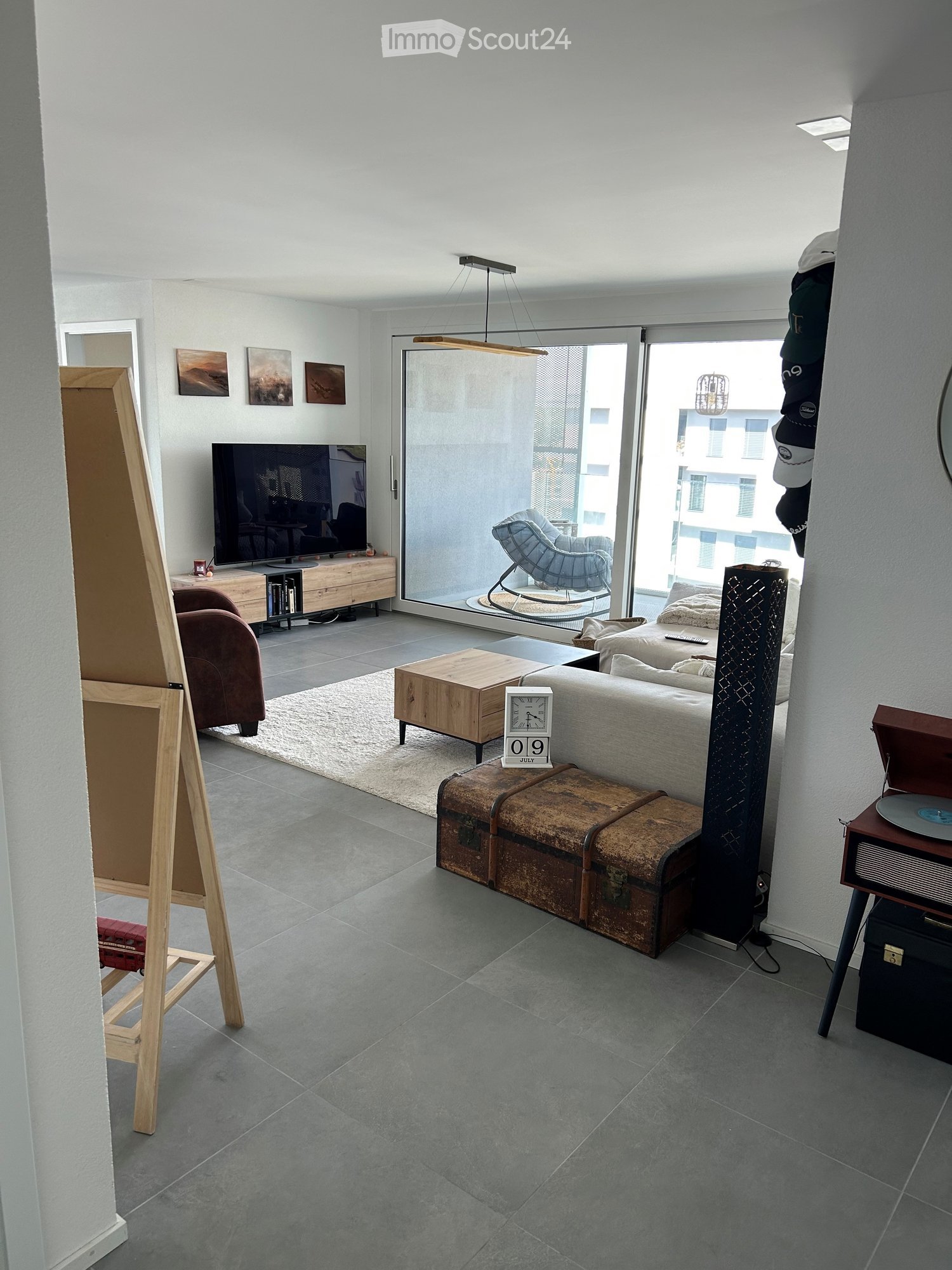 Modern living room with grey sofa, coffee table, armchair, TV, wooden cabinet, clock, trunk, and record player. Large windows and a view of the city.