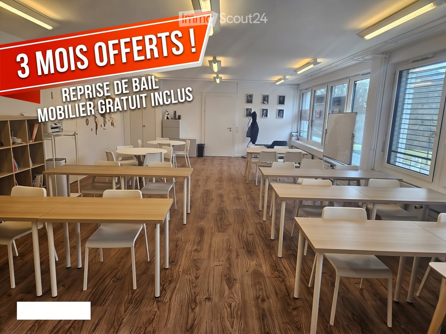 Bright, modern room with wooden flooring, multiple tables and chairs arranged for a collaborative workspace, shelves, a refrigerator, a printer, and a whiteboard.