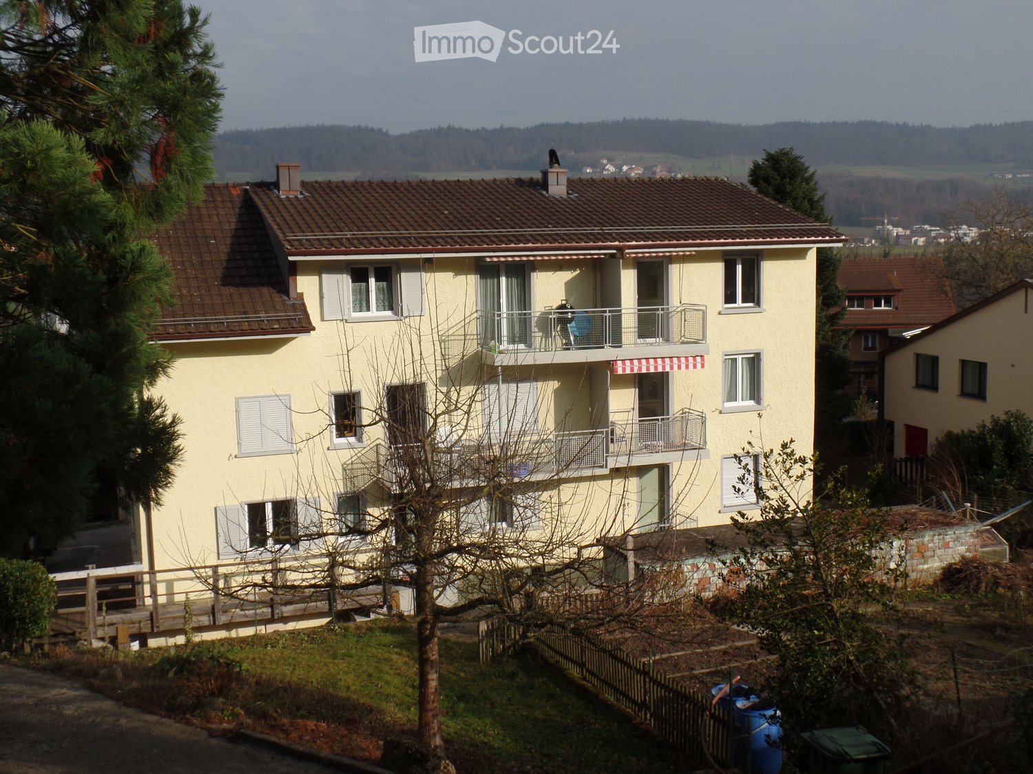 Two-story yellow house with brown tiled roof, multiple windows, balconies, and surrounding greenery.