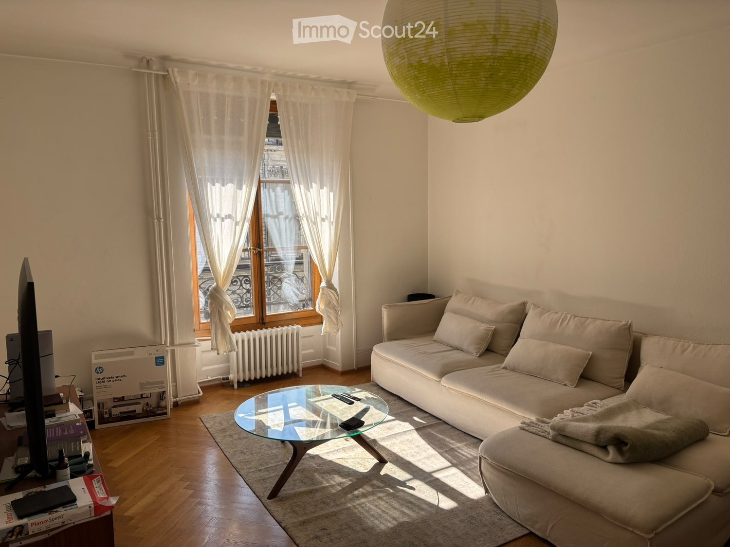 Bright living room with a white sofa, glass coffee table, TV, and a large window with sheer curtains. The room features wooden flooring and a decorative balloon lamp.