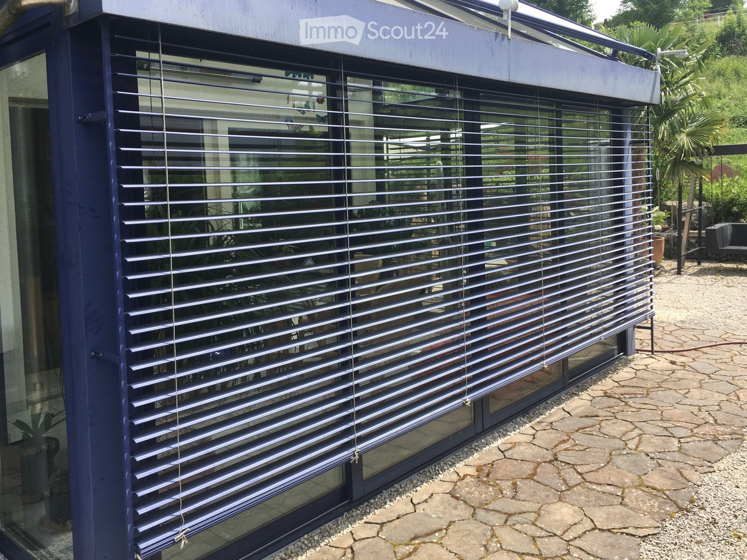 A modern balcony or terrace with blue roller blinds, stone pavement, and surrounding greenery.