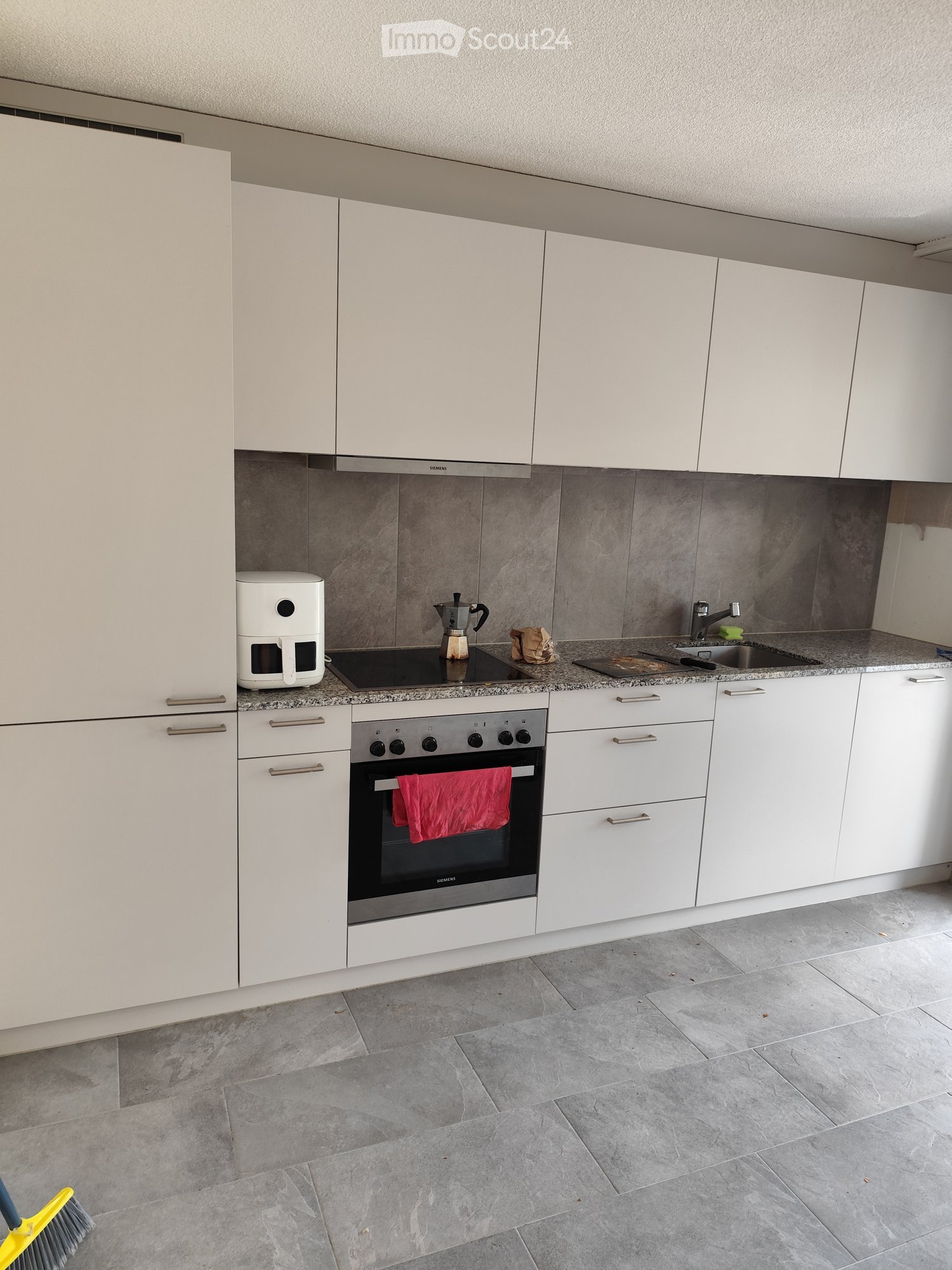 Modern kitchen with white cabinets, granite countertops, and stainless steel appliances including an oven, coffee maker, and induction stove. A red towel hangs from the oven door.