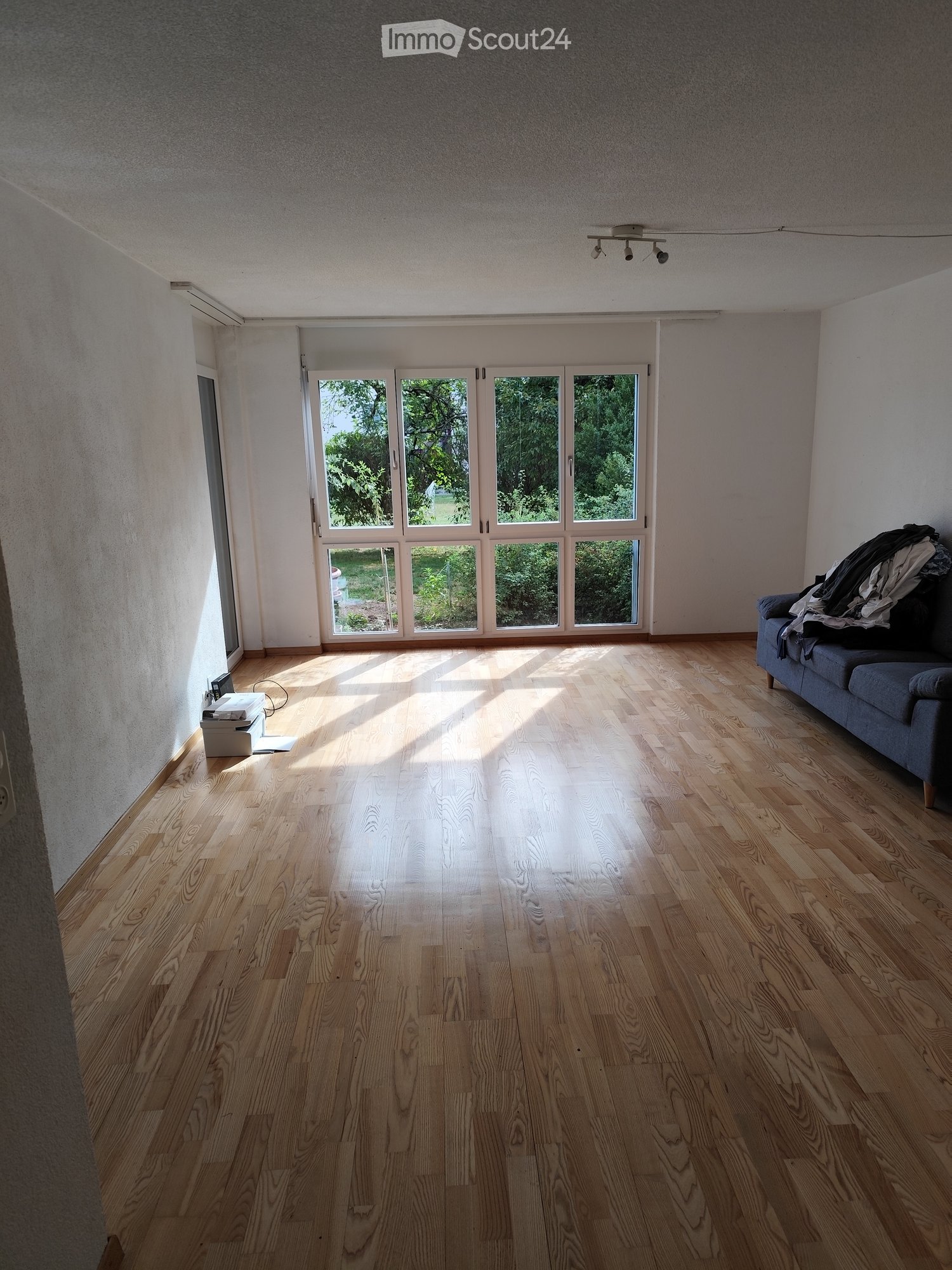 Bright, empty living room with wooden flooring, large windows, and a sofa with some items on it.