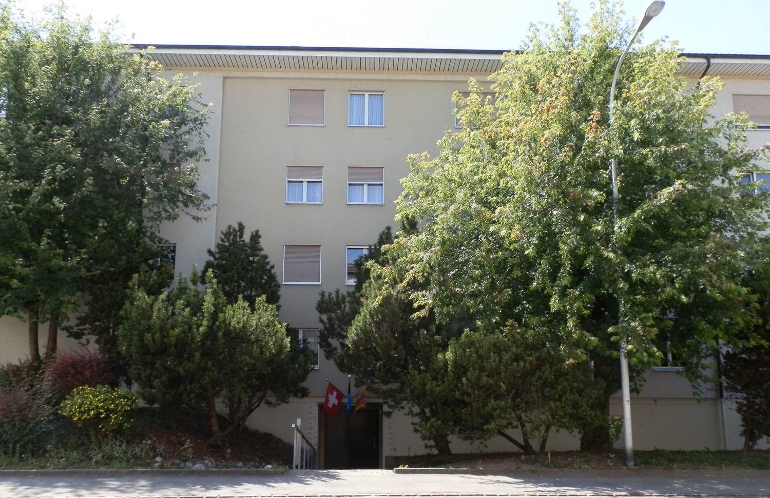 A multi-story residential building with beige walls, multiple windows, and a small entrance area surrounded by trees and greenery.