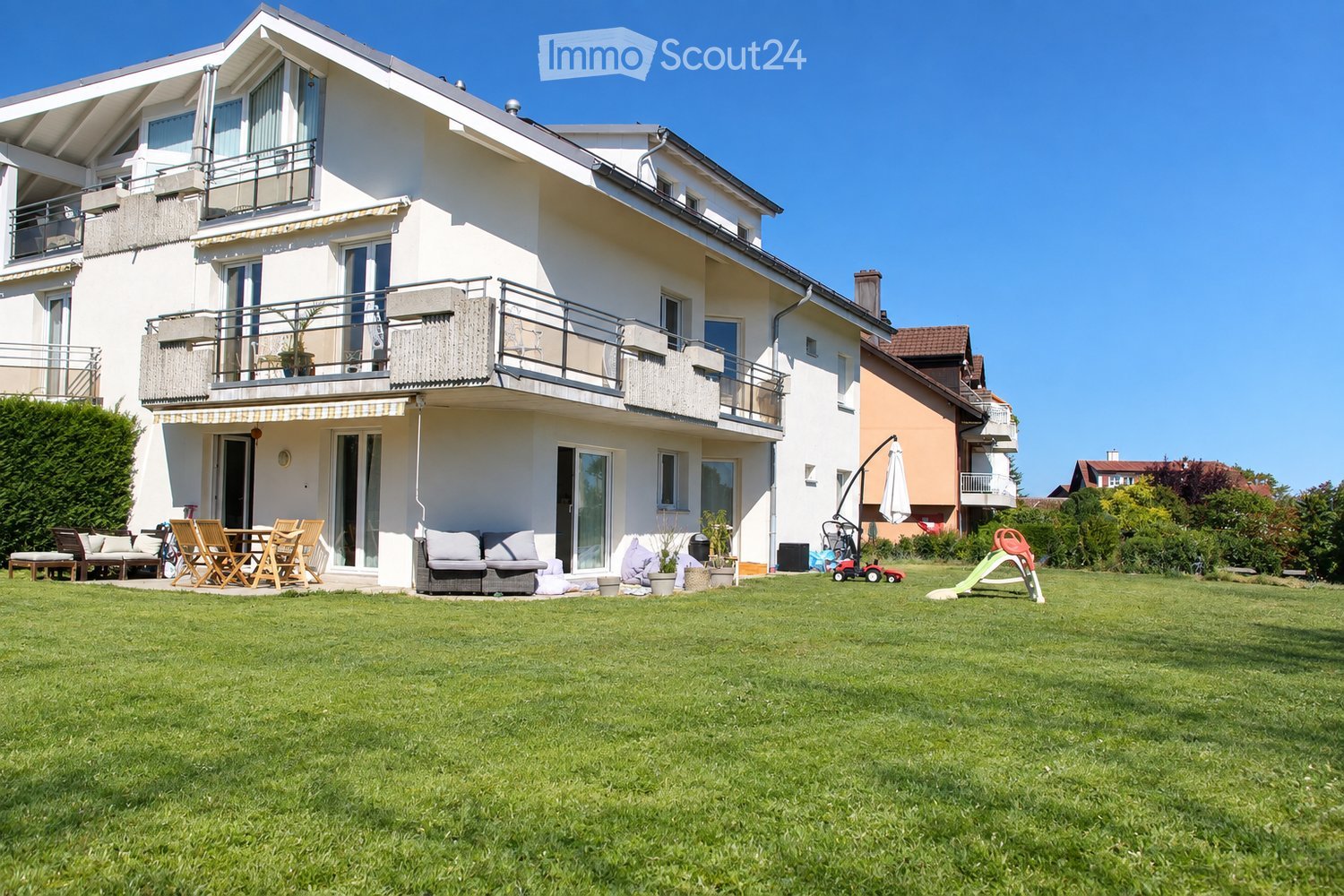 Two-story house with white exterior, multiple balconies, large windows, and a well-maintained lawn with outdoor furniture and play equipment.