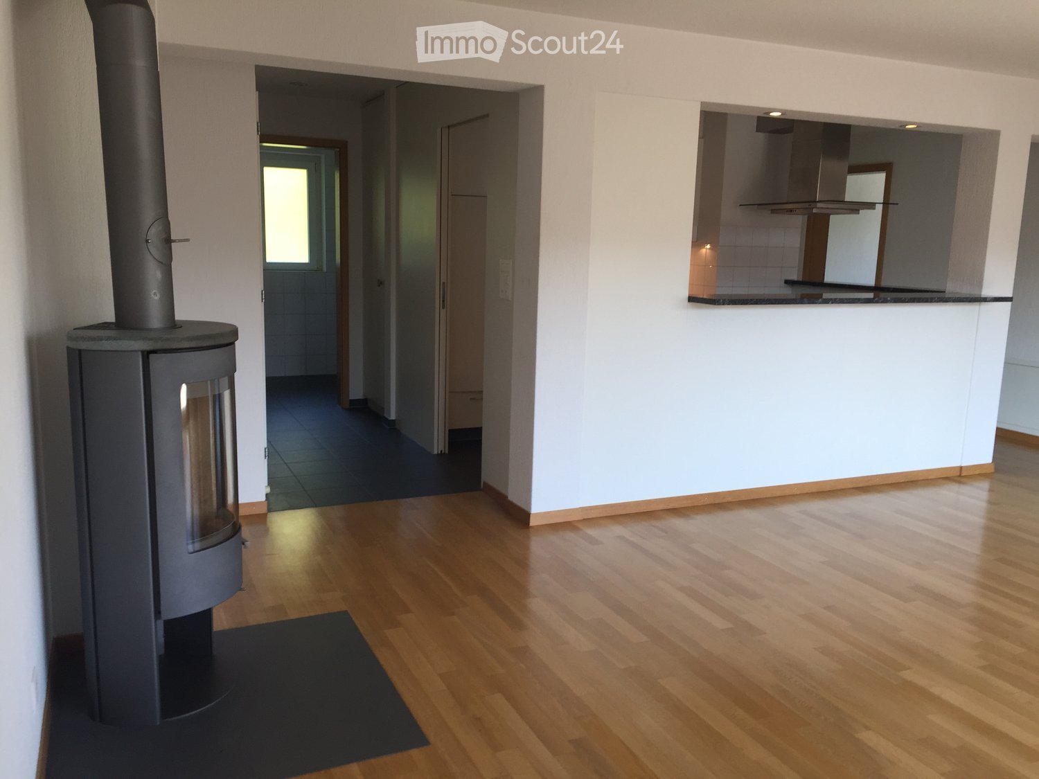 Bright hallway with wooden flooring, white walls, and a modern wood-burning stove on the left. The hallway leads to a kitchen area visible on the right with white cabinets and a countertop.