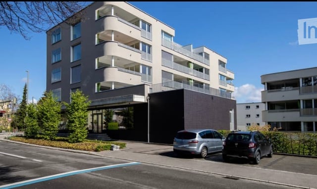 Modern multi-story residential building with balconies and a covered entrance, two parked cars in front.