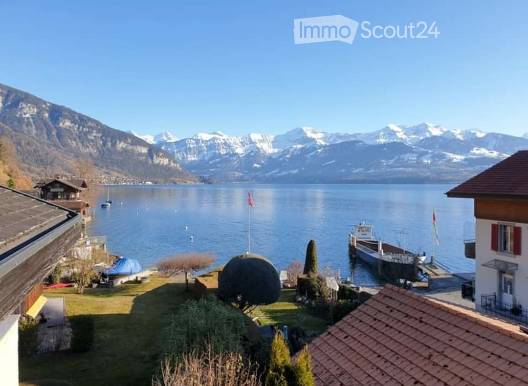 View of a lake with snow-capped mountains in the background, featuring a well-maintained garden area with trees, a flagpole, and a dock with boats.
