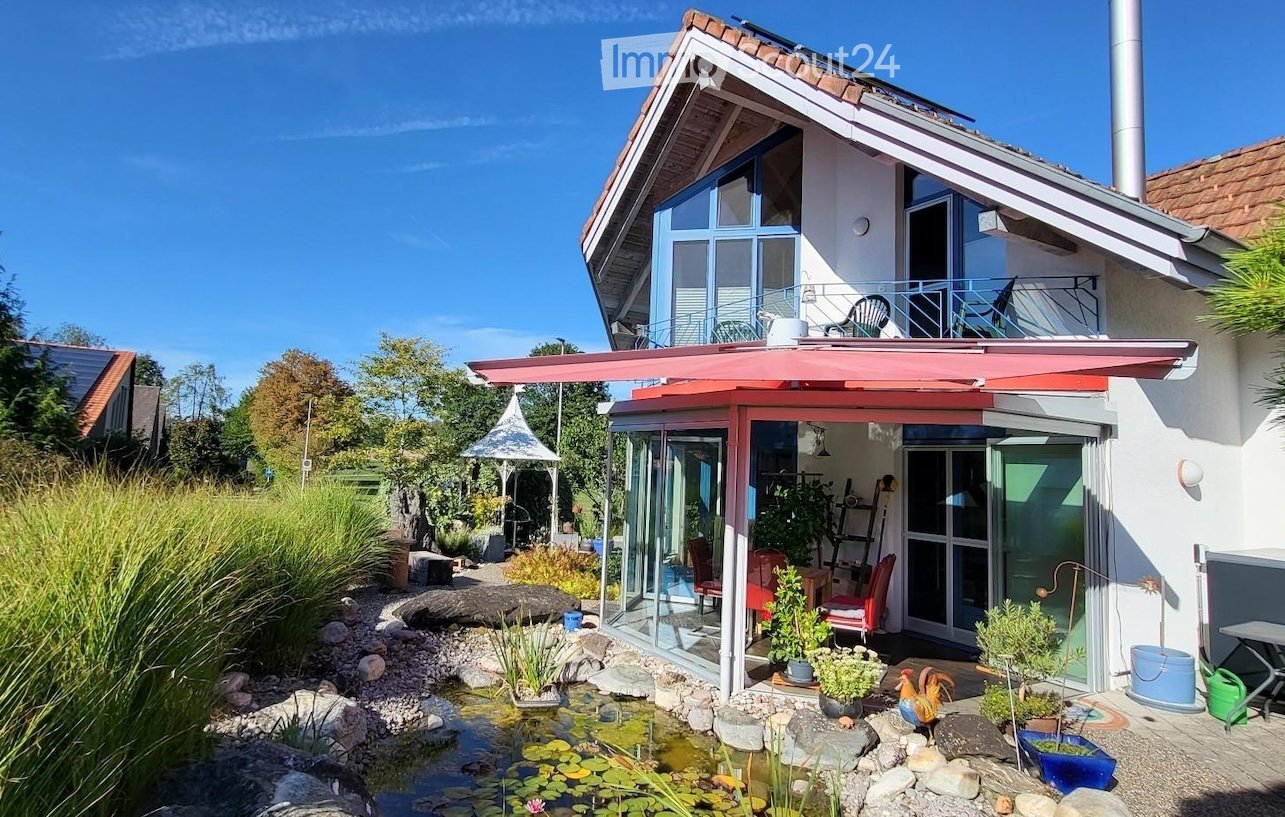 Modern two-story house with a red roof, large windows, and a spacious garden area featuring a pond, plants, and outdoor furniture.