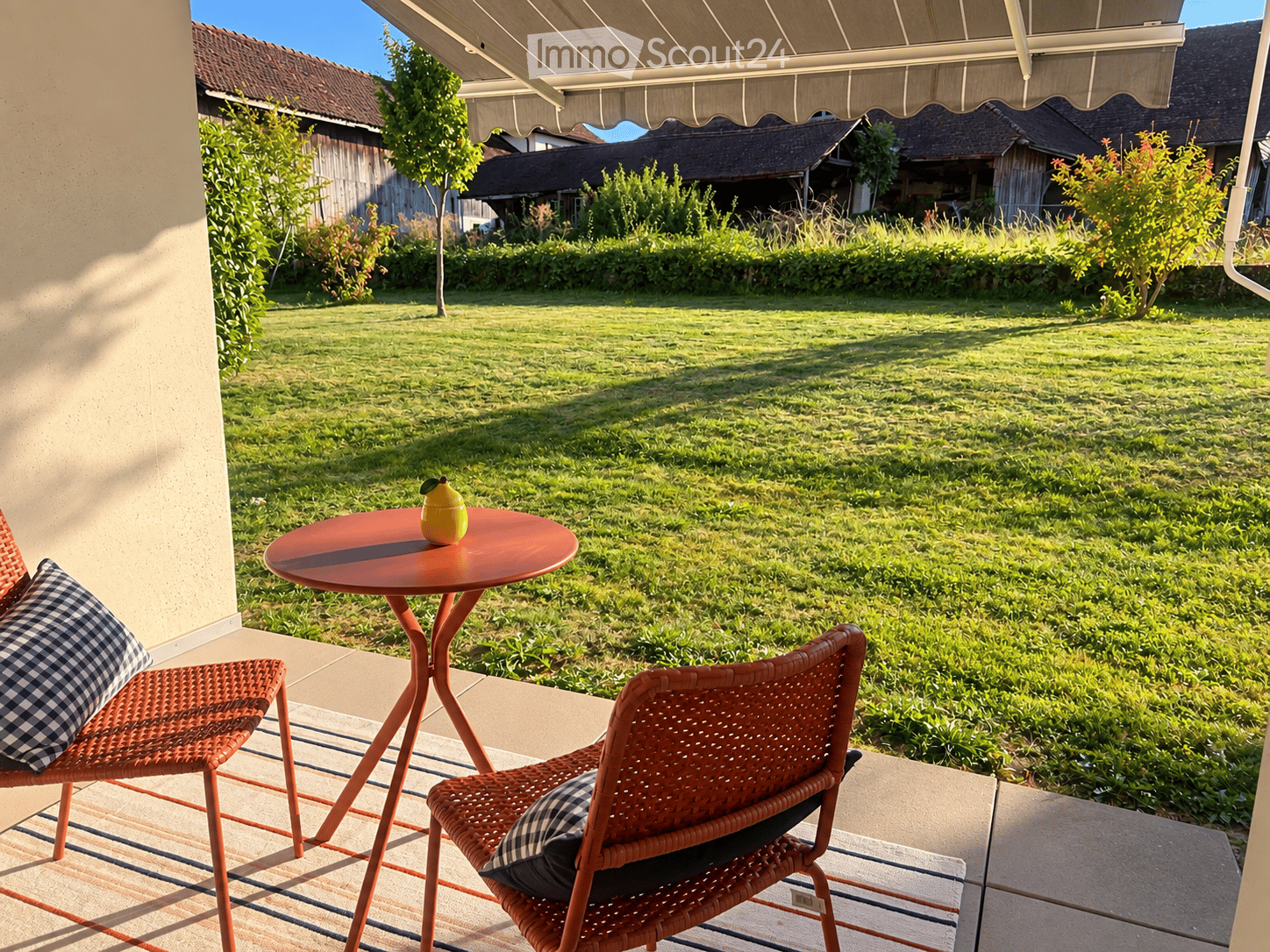 A sunny outdoor patio with a round table, two wicker chairs, and a small fruit bowl, overlooking a well-maintained lawn and garden area.