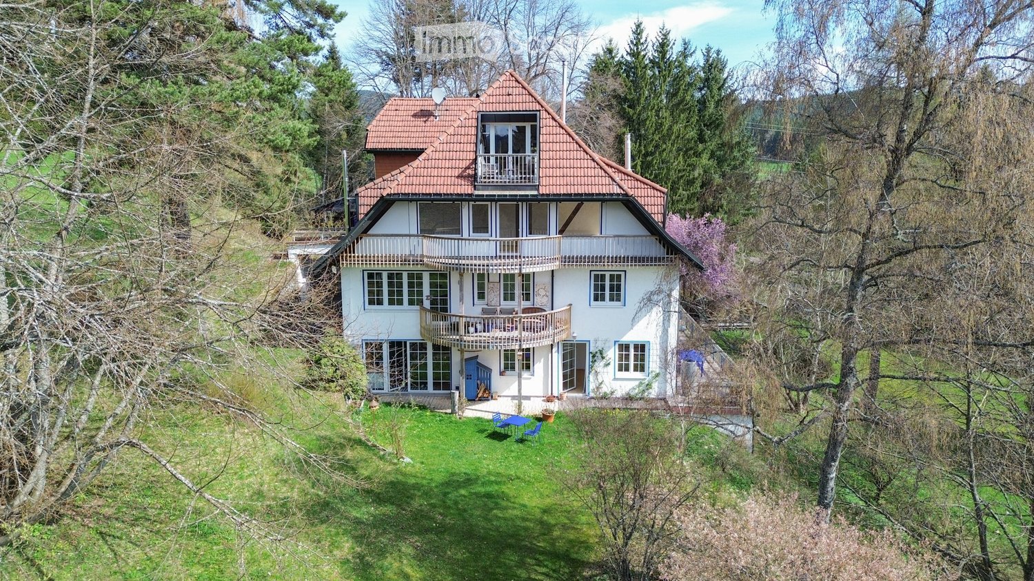 Two-story house with red-tiled roof, white walls, multiple windows, balconies, and a small garden area with outdoor furniture.