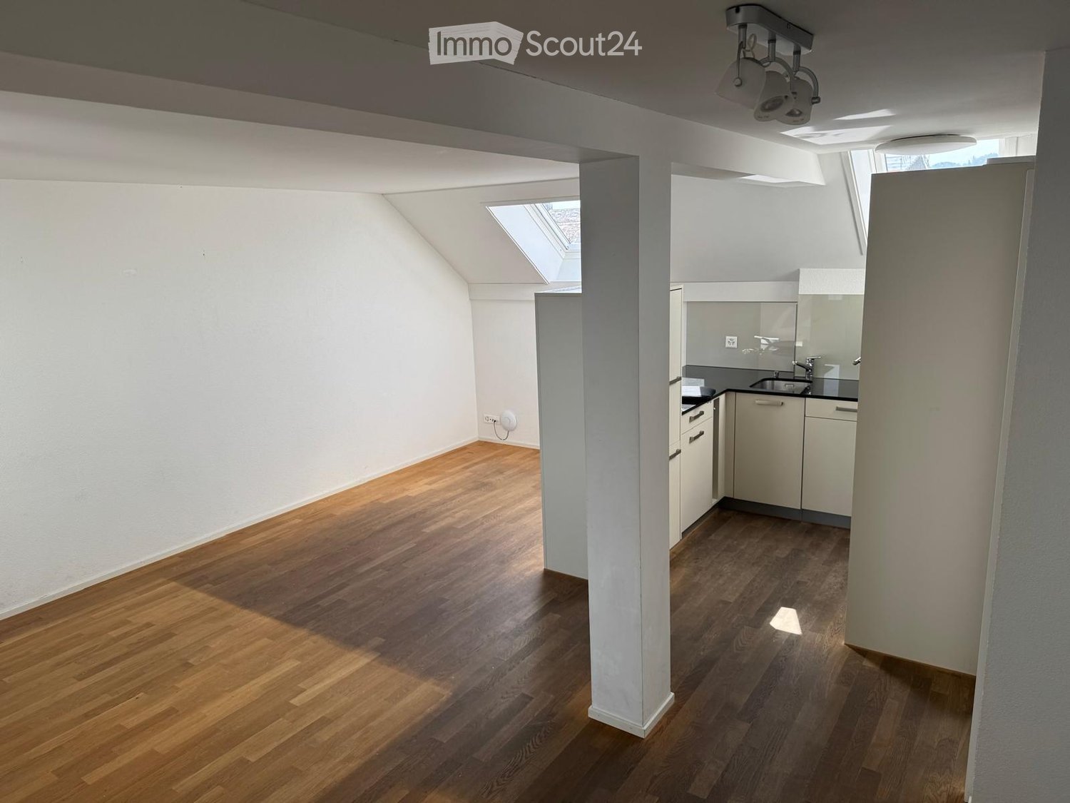 Bright, open loft-style room with wooden flooring, white walls, and a small kitchenette area featuring white cabinets and a sink.