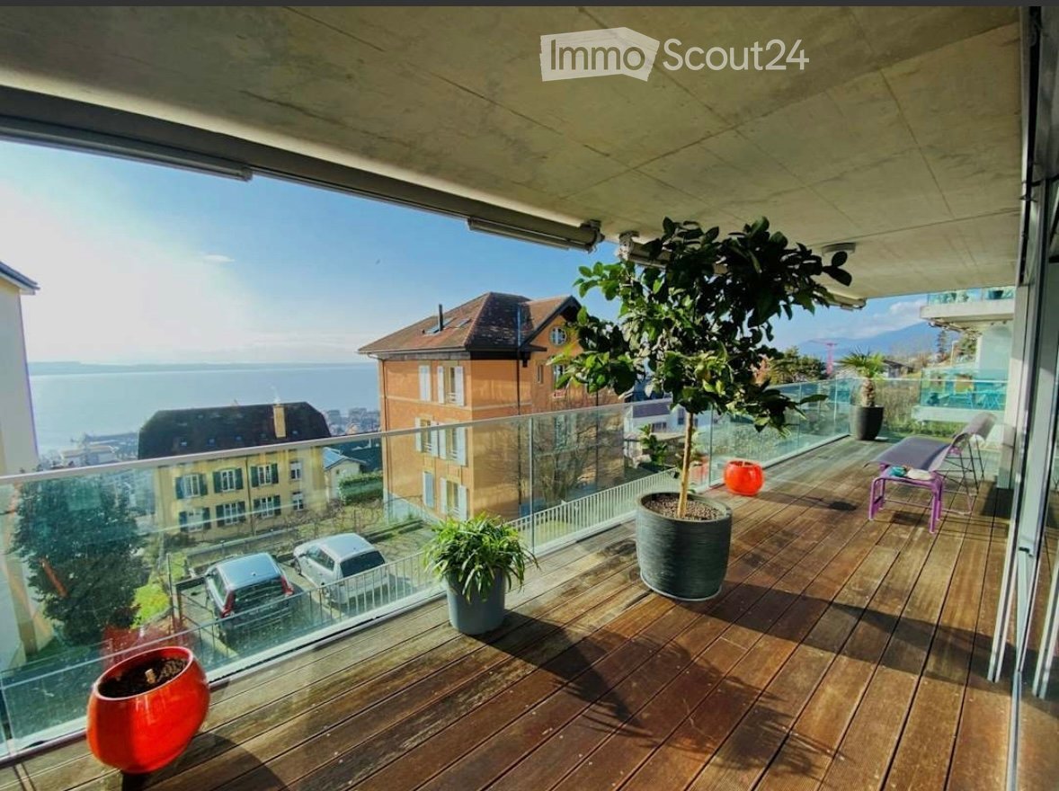 Balcony with wooden decking, glass railing, potted plants, and a view of the sea and surrounding buildings.