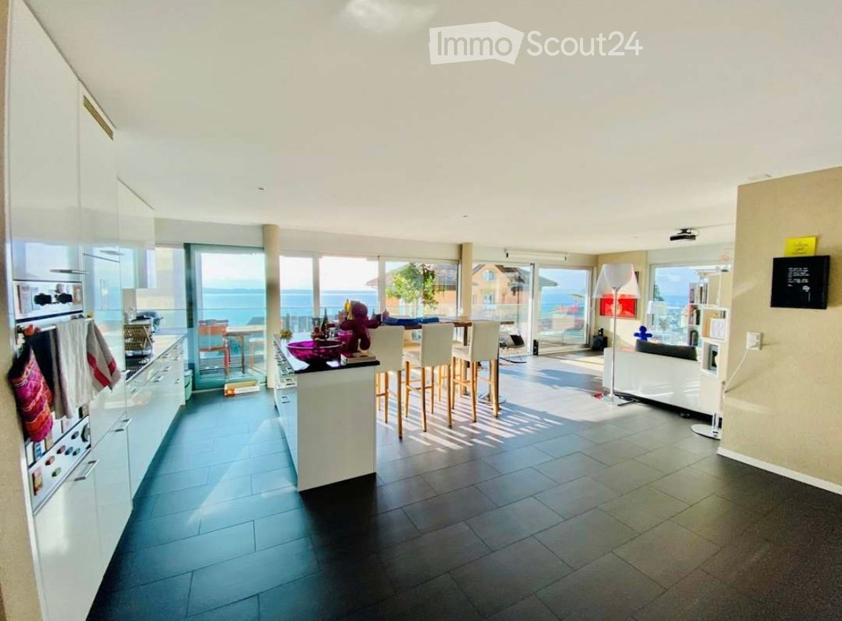 Modern kitchen and living area with large windows offering a sea view, equipped with a refrigerator, oven, kitchen island, bar stools, sofa, and floor lamp.