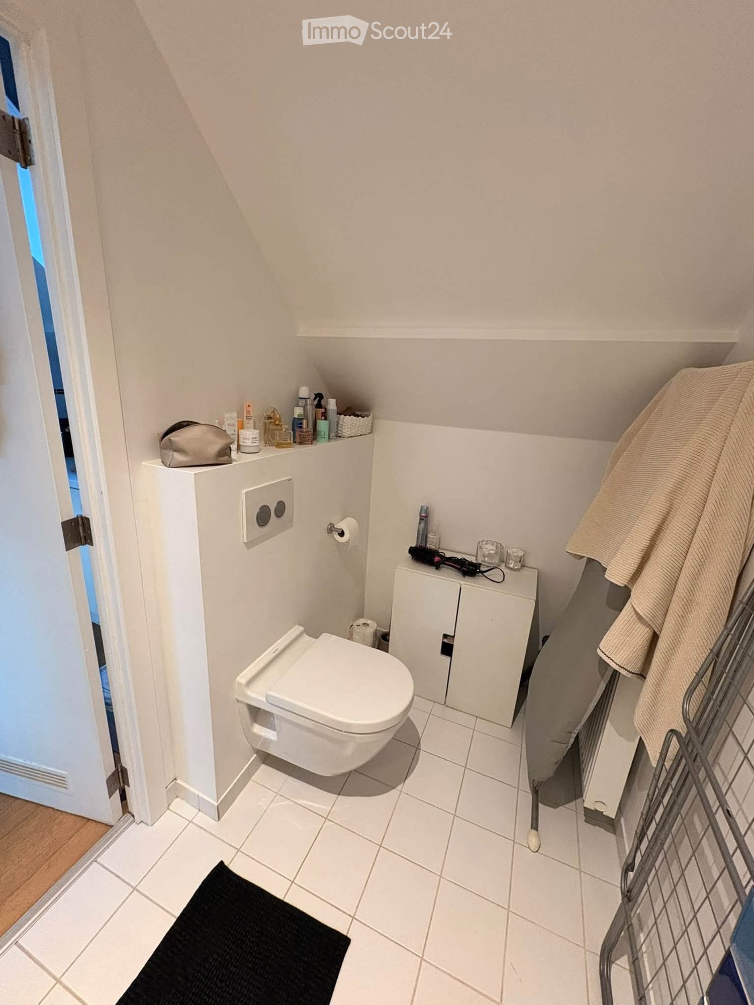Compact bathroom with white tiled floor and walls, toilet, small cabinet, mirror, towel, and personal care items on shelves.