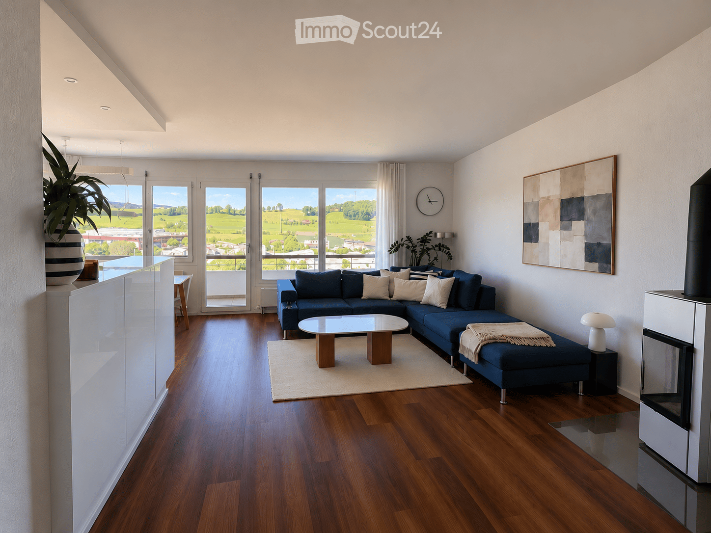 Modern living room with wooden flooring, large windows offering a scenic view, a blue sectional sofa, a glass coffee table, a fireplace, and decorative artwork.