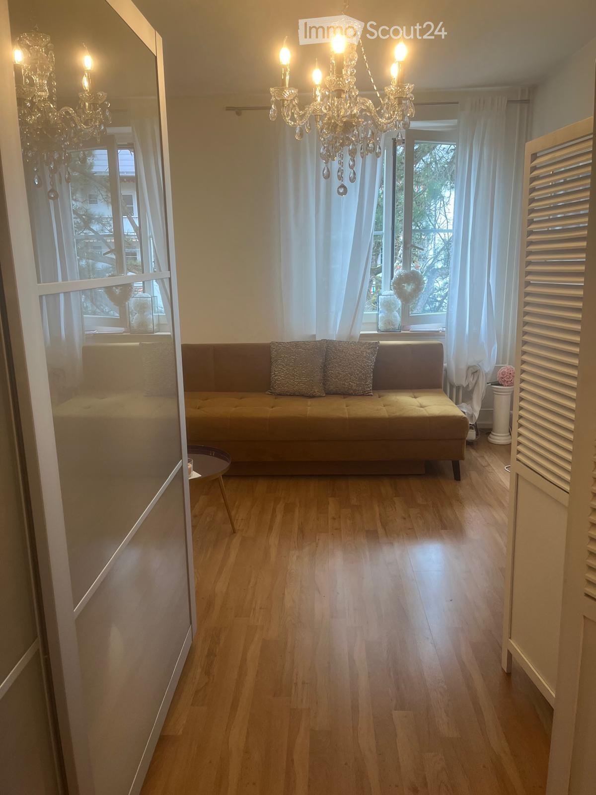 A well-lit living room with wooden flooring, a beige sofa with patterned cushions, a small round table, and two chandeliers hanging from the ceiling. Large windows with white curtains allow natural light to fill the space.