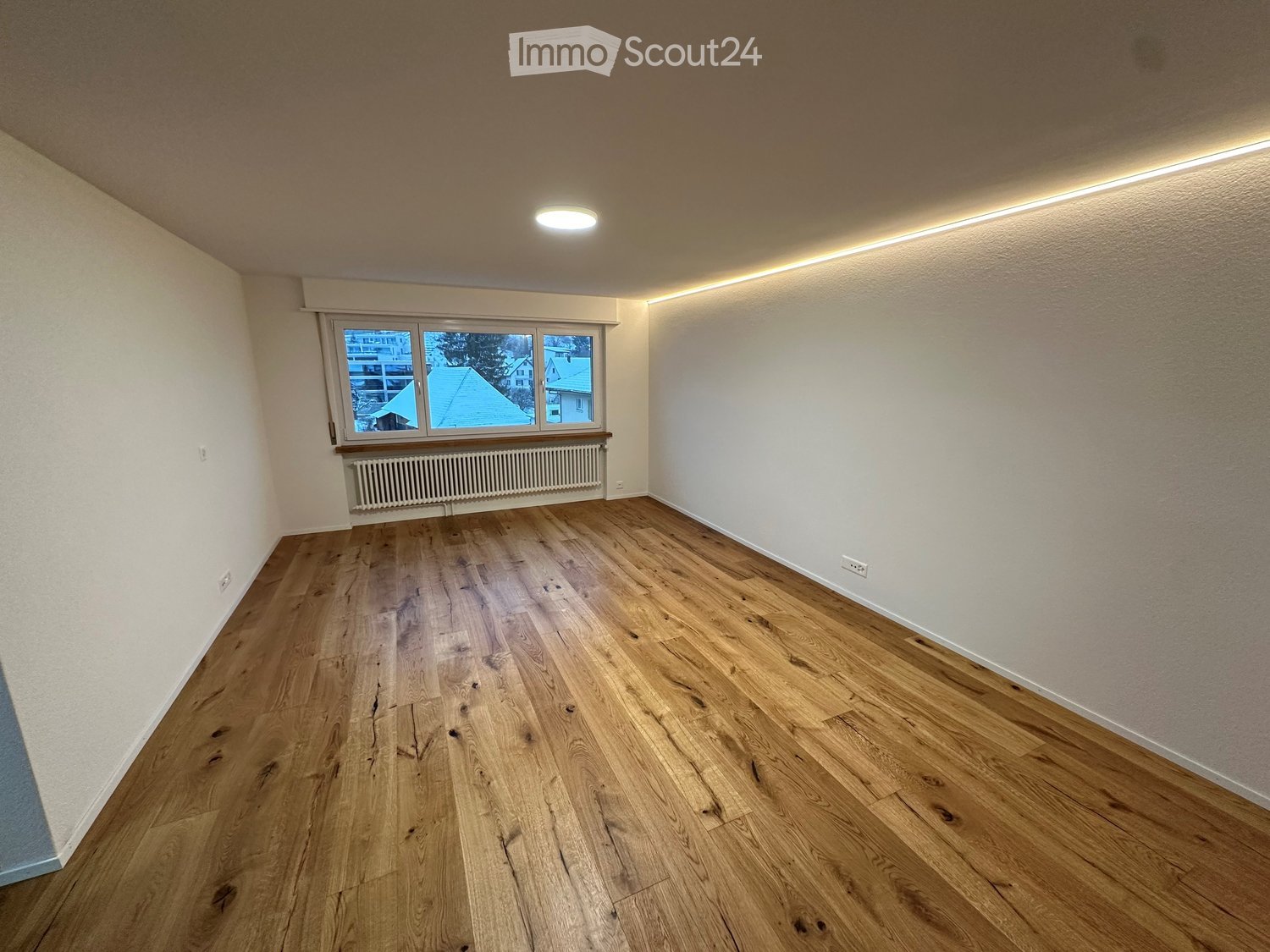 Empty room with wooden flooring, white walls, large window, radiator, and recessed lighting.