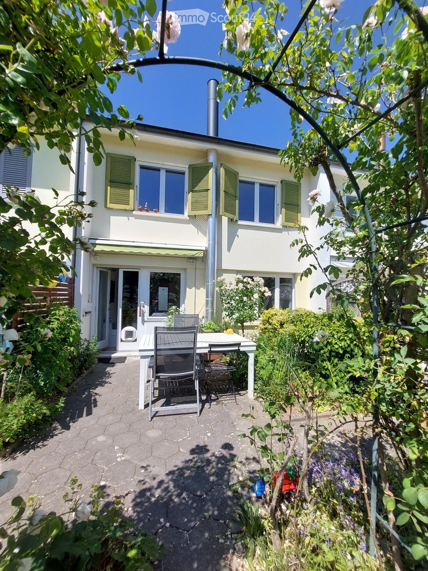 A well-maintained garden area with a table and chairs, surrounded by lush greenery and a two-story house with green shutters and a chimney.