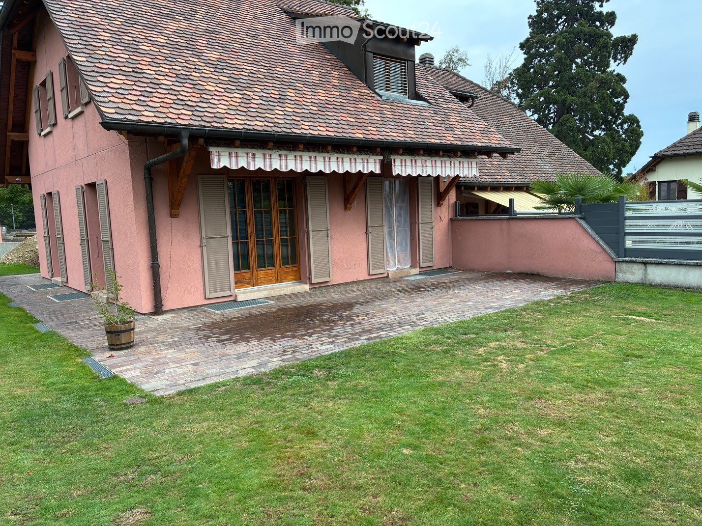 Pink two-story house with a tiled roof, wooden shutters, and a covered entrance. The house is surrounded by a well-maintained lawn and a paved driveway.
