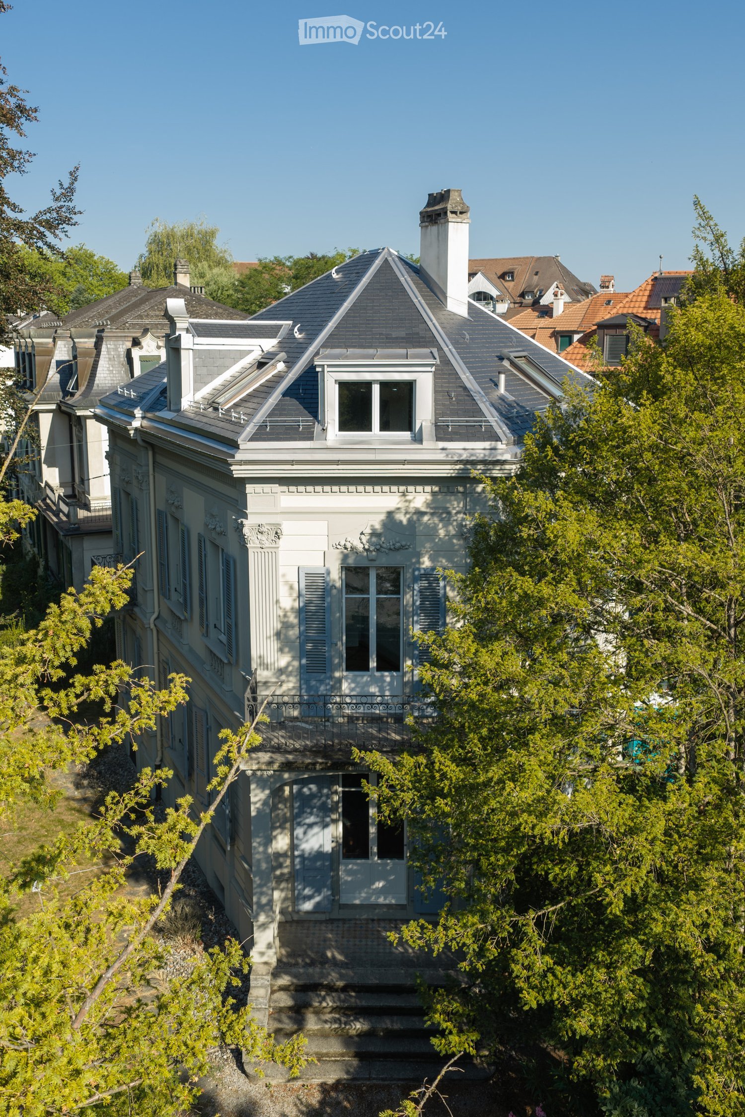A multi-story house with a grey roof, white walls, and ornate architectural details, surrounded by trees and greenery.