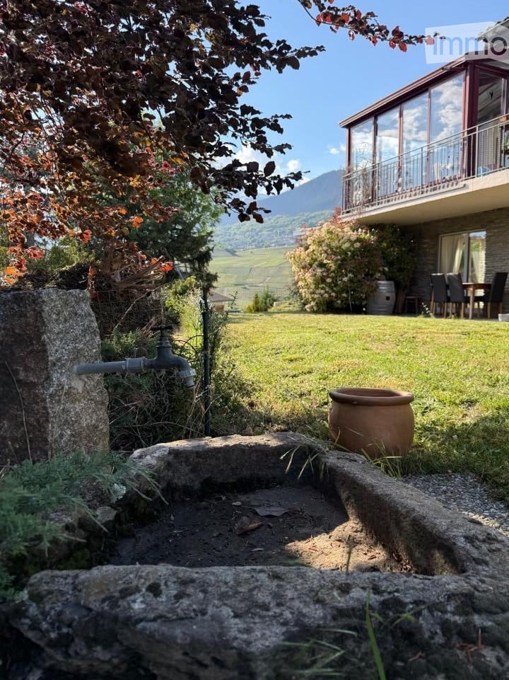 Lush garden with a stone well, outdoor faucet, and a large ceramic pot. The background features a modern house with a balcony overlooking a scenic valley.
