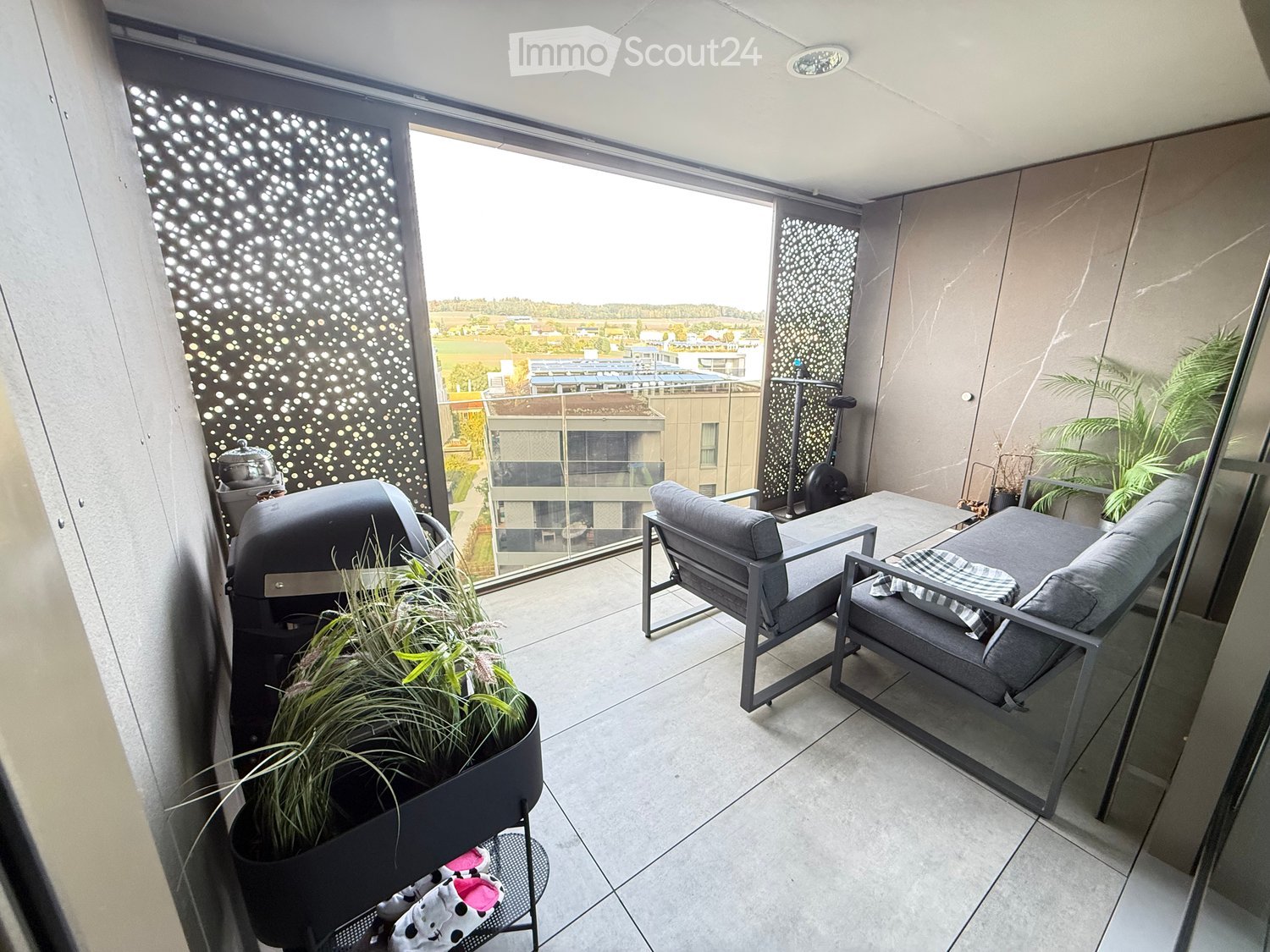 Modern balcony with grey furniture, including a sofa, chairs, a small table, and a barbecue. Features a decorative screen, potted plants, and a view of the surrounding area.