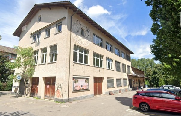 Two-story building with beige facade, multiple windows, and a mural on the side. There is a parking area with a red car in front.