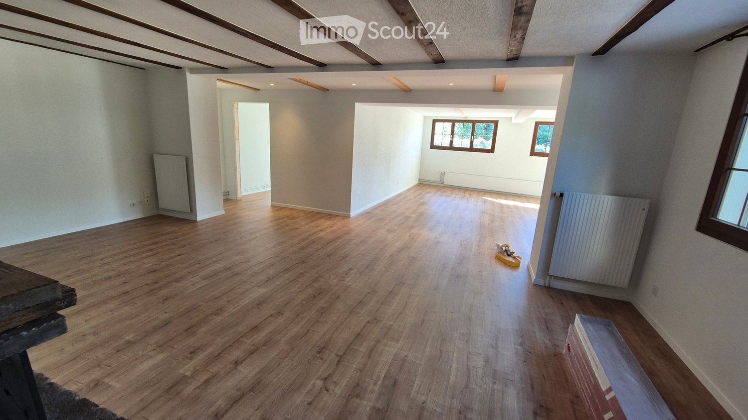 Empty room with wooden flooring, white walls, exposed wooden beams on the ceiling, and natural light from windows. Radiators are visible on the walls.